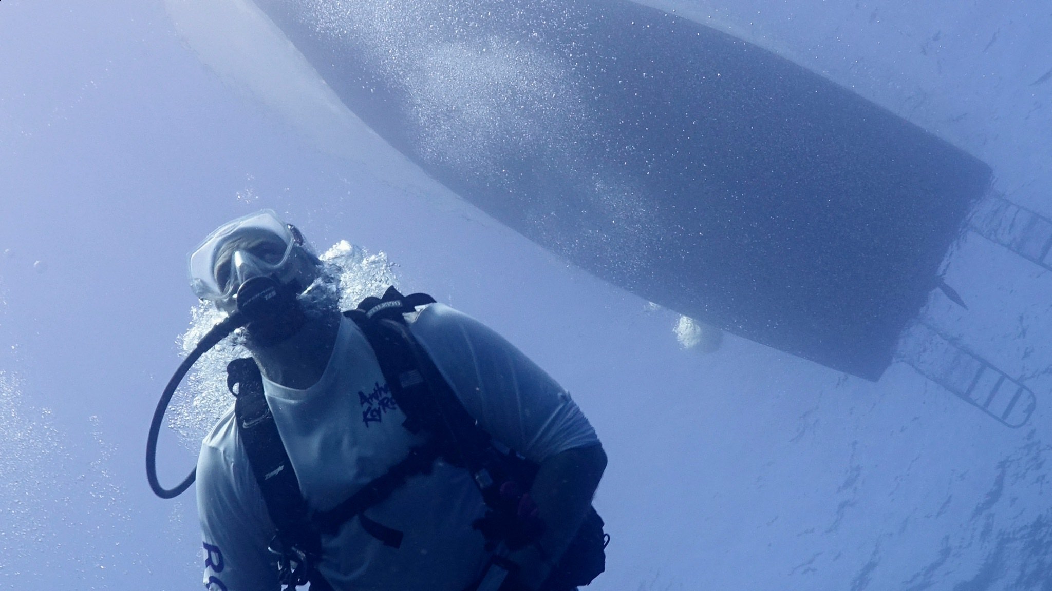 A scuba diver hovering beside the dive boat's hull, seen from below with sunlight streaming through the surface